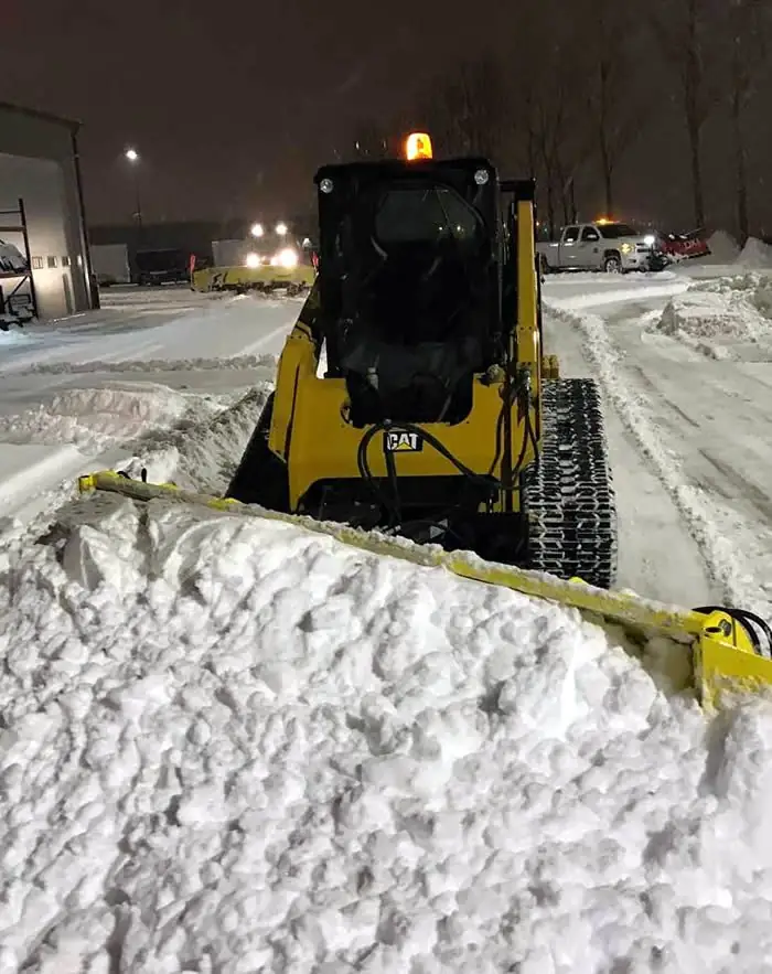 Tracked skid steer with a snow pusher stacking snow in a commercial lot at night